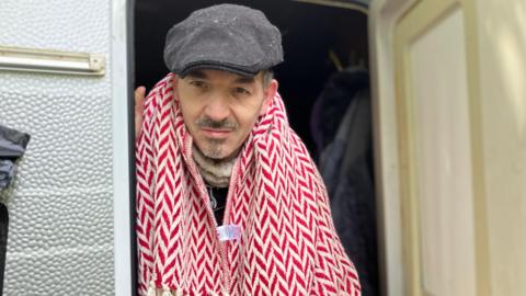 Dan Ampleford. He is pictured standing in the door of a caravan, wearing a grey hat and a red and white patterned scarf. He has a small amount of grey facial hair. 