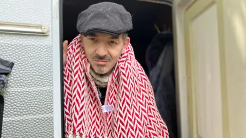 BBC Dan Ampleford. He is pictured standing in the door of a caravan, wearing a grey hat and a red and white patterned scarf. He has a small amount of grey facial hair. 