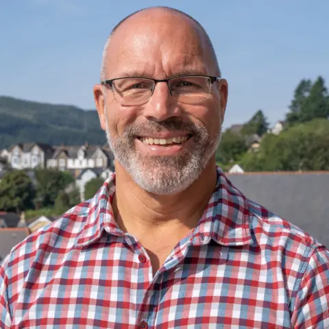 TFW Lee, a balding man smiles at the camera. He is wearing a red, white and blue check shirt and has short grey stubble. He has thin-rimmed classes on and behind him houses and trees can be seen. 