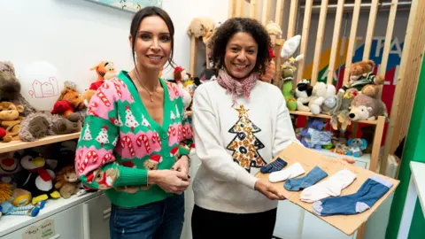 Little Village Christine Lampard and Renata Acioli stand at the charity's hub, surrounded by donations and are both wearing Christmas jumpers 