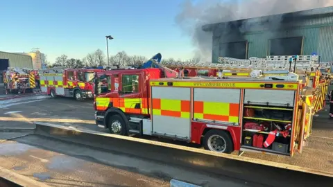 At least four fire engines parked up in front of a warehouse. Black smoke is billowing from the building into a light blue sky. 
