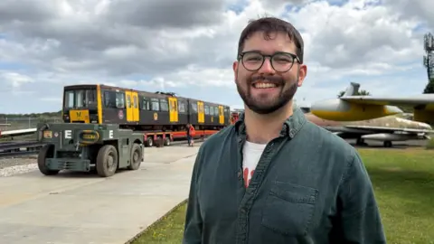 Adam Dixon is smiling, as the original black and yellow Metro train sits in the distance. Dxon has short brown hair and beard and circular black-rimmed glasses. He is wearing a grey buttoned shirt and a white t-shirt.