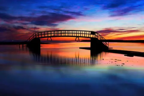 John Cuthbert Sunrise sky of purple, blue, orange and yellow, reflected in water, with a bridge across the water.