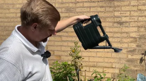 Amber Ward/BBC A man in a white T shirt pours water on to plants using a black watering can