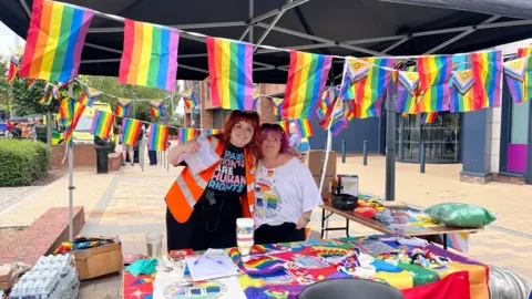 Two women stood under a black canopy with Pride flags hanging from it in front and behind them. The woman on the left is organiser of the event, Elise Gilbert. She is wearing black trousers, a black top with blue and pink bold writing on and an orange hi-visibility vest on. She has short red hair with a fringe and has blue, yellow and orange stripes pained on both of her cheeks. She has her arm around a woman with short pink hair and a fringe. She is wearing a white t-shirt with a colourful logo on and black trousers. There are tables with colourful decorations on in front of them and to their left.