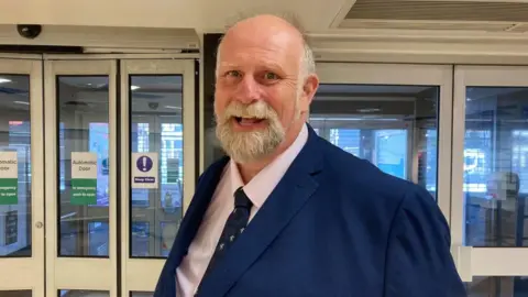 Alex Pope/BBC Tom Wootton, the mayor of Bedford, wearing a blue suit, standing in front of doors inside the empty former Debenhams store in Bedford 