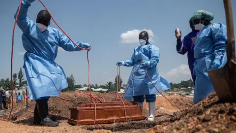 AFP via Getty Images Four people wearing personal protective gear lower a coffin into a grave using ropes