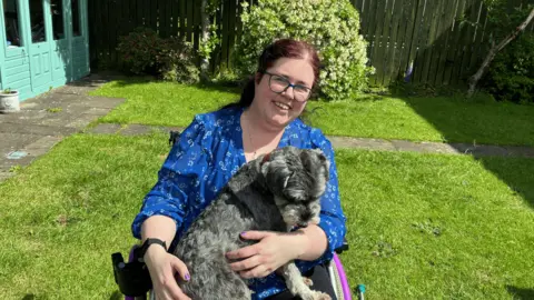 Victoria Gerrard A woman with dark red hair, pulled back into a ponytail, sits in a wheelchair in a sunny garden, with a grey-haired dog on her lap. She is smiling at the camera and wears a blue blouse with a floral print.