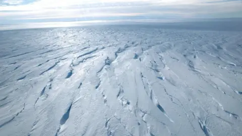 National Environment Research Council via PA View of Thwaites Glacier which extends into the distance as a white, crevassed sheet of ice. The sky is light blue with cloud cover.