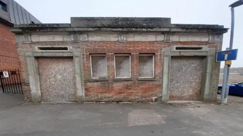 An exterior shot showing the front of the empty red-brick toilet block which has boarded windows and doors.