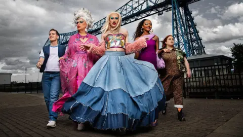 The Smoggie Queens cast lined up walking towards the camera with an industrial structure behind them.