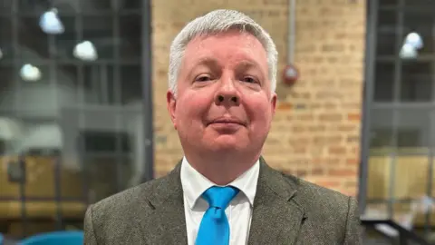 Peterborough City Council Andrew O'Neil looking into camera in the council chamber. He has short white hair and is wearing a grey suit, white shirt and blue tie.