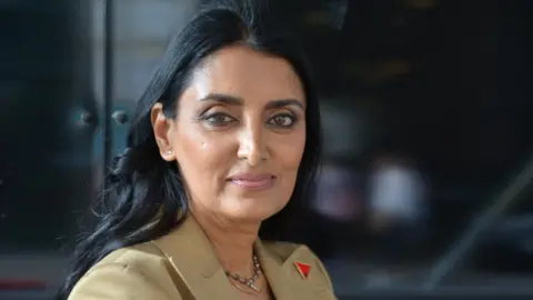 Aneeta Prem Aneeta Prem looks directly at the camera. She has dark hair and is wearing a pale coloured suit jacket. There is a red and black badge on her lapel in the shape of a triangle. 