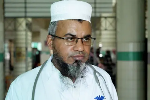 Close-up of a doctor standing in a hospital corridor in Mymensingh, Bangladesh, wearing glasses, a white cap, and a white coat with a stethoscope around his neck
