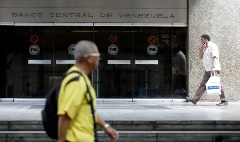 Marco Bello / Reuters A person wearing a yellow T shirt and a backpack walks past the central bank of Venezuela as another person in a white shirt walks holding a shopping bag in the background, outside a glass entrance with the words 'Banco Central de Venezuela' on it, in Caracas in 2017.