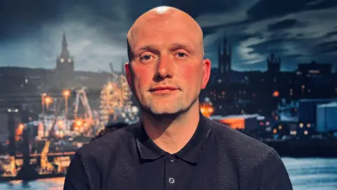 Politician Stephen Flynn in front of an image of Aberdeen harbour, looking at the camera.