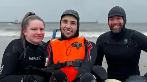 Left to right, Lauryn Bell, Jimmy Simpson and Nick Jones are at the edge of the sea. All are wearing black wetsuits and Mr Simpson also has on an orange lifesaver. He and Mr Jones also have on peaked wetsuit-style head coverings. Ms Bell has her long dark blonde hair - which looks brown in the wet - tied up in a high ponytail.