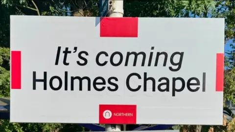 Railway station sign reading: 'It's coming Holmes Chapel'.
