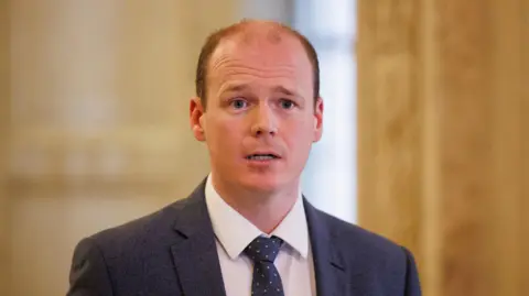 A man with short red/brown hair looks to the right of the camera. He wears a dark grey suit, white shirt and Navy tie. Behind him is an out of focus dark cream wall. 
