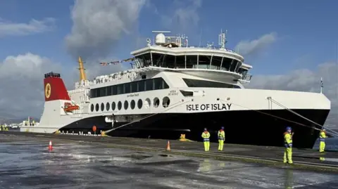 Brian Fulton A black and white ferry on water with Caledonian MacBrayne written on it, with Isle of Islay written near the front. It is docked and workers in fluorescent gear stand by it