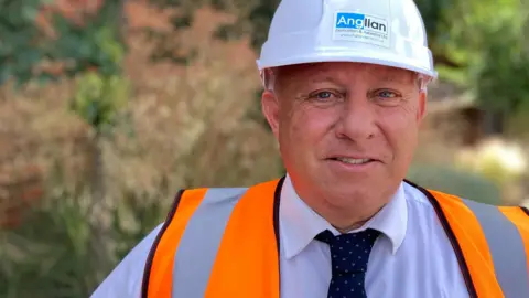 Lee Storer in a hard hat and a high-viz jacket. He is wearing a blue shirt and blue-spotted tie. The background is out of focus.