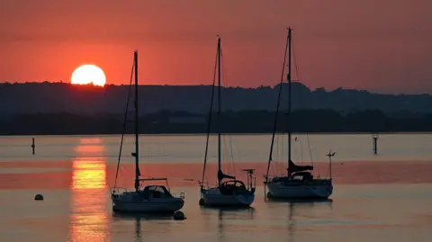 Alison Treacher The sun rises above the hills and casts it's early orange hues over the water and across the hazy skies. Three moored small yachts can be seen on floating through the middle of the shot with their sales safely stowed.