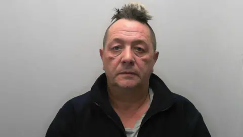 A middle aged man wearing a black fleece jacket stares forwards in a police mugshot. He has a short untidy Mohican-style haircut.