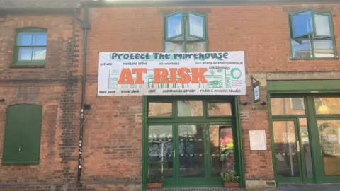 Exterior of a red-brick building with green-framed windows and doors. A large banner reading “Protect The Warehouse - At Risk” hangs above the entrance, promoting a community garden, bike shop, book shop, meeting space, co-working, radio and podcast studio, workshops, and environmental initiatives. 