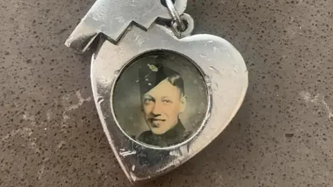 Claire Stonier A heart-shaped silver pendant on a pin with a circular frame containing an old black and white photograph of a man smiling and wearing a garrison cap with a military badge on the side