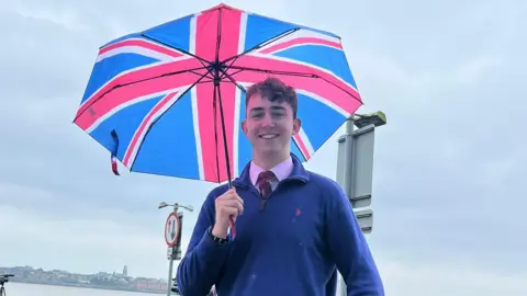 supplied Harry smiles at the camera, he is wearing a blue sweater with a pink shirt and burgundy tie. He is holding an umbrella with a union jack print on it.