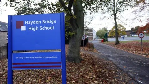 A blue sign with white lettering for Haydon Bridge High School on a patch of grass covered with brown leaves. It is placed in front of a tree. To the right is a path leading to the school.