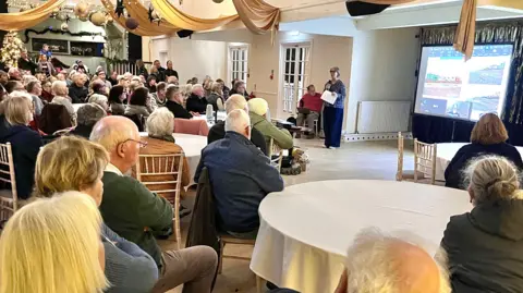 Martin Giles/BBC Tens of people sits in chairs behind circular tables as they look and listen to a woman presenting at the front of a hall. The woman is speaking into a microphone with a large projection board next to her.