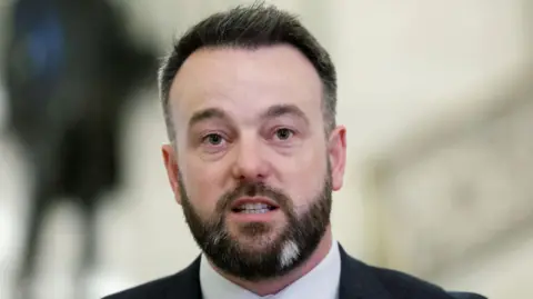 Colum Eastwood - a man with dark hair and beard wearing a suit looks beyond the camera
