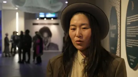 Qian is standing inside the museum's building close to a wall with a photo of her husband behind her in the distance. She is wearing a brown hat and has long, brown hair and a brown jacket on.