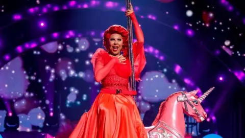 A performer on Strictly is wearing a red dress, has red hair and is sitting on a pink unicorn.