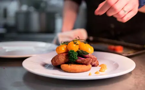 A person's hand can be seen hovering above a nicely plated dish of steak, tomatoes and other vegetables in an industrial kitchen. 