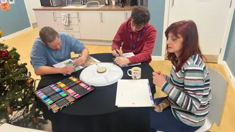 Three people, a woman and two men, sit around a table writing and colouring in a kitchen, with a Christmas tree in the corner