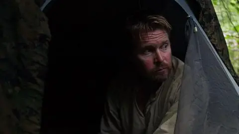 A man with a short light brown beard sits in a tent pitched in a forest clearing. He looks lost in thought.