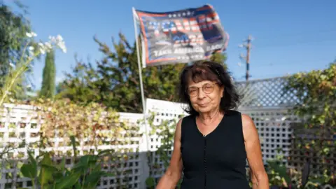 Philippe Studio June in her garden, looking at the camera smiling, in front of a white fence with plants, a blue sky and a flag printed with the words Trump 2024.  She is wearing a black cropped top and round wireframe glasses.