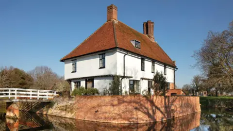 A general view of Letheringham Lodge - a white building with an orange tiled roof surrounded by a moat and trees. 
