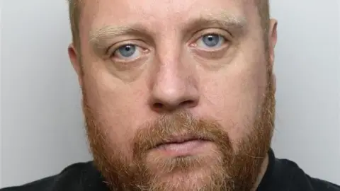 A custody picture of a man with auburn hair and beard. He is wearing a black top.