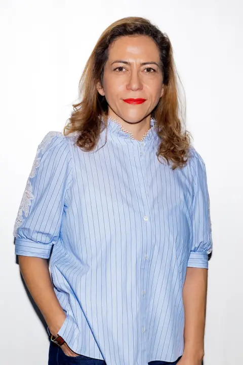 Soudade Kaadan A woman with long dark-blonde hair looks at the camera She has on bright red lipstick and is wearing a blue shirt with rolled-up sleeves
