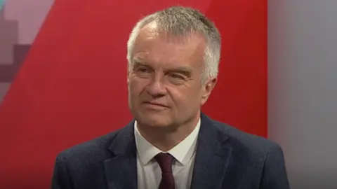 A man with short grey hair stands on a pavement with parked cars on the road. He is wearing an open-collared shirt under a brown jacket.