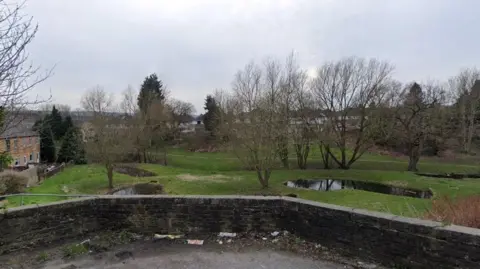 Google Maps The picture overlooks a green space with several trees and hedges. Two small bodies of water can be seen. To the left, a row of houses stretches.