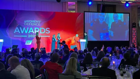 Royal Navy Wide shot of 2025 Women in Defence Awards ceremony showing crowd and stage, with a surprised Abigail Wright visible on a large screen