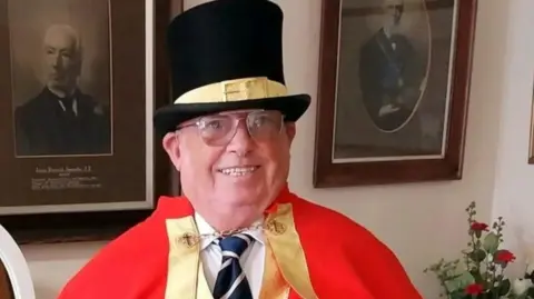 Terrence Mullett A man wearing a black top hat with a yellow buckled ribbon across the middle and is wearing a red cloak over a white shirt and blue striped tie with a golden clasp fashioning the cloak.