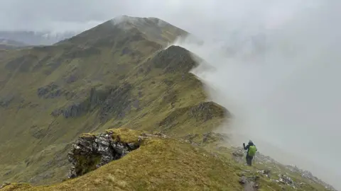 JEANNIE MCLEAN A mountain ridge covered in grass with a hiker wearing dark clothing and a light green backpack walking with poles in the foreground. Part of the left side of the image is covered with a thick white-grey fog. The sky is overcast.