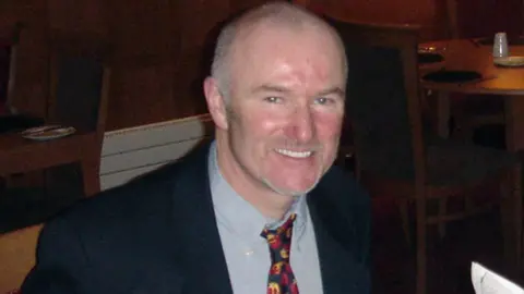 Dr Mohammad Husban Colin Howell is smiling at the camera. His hair is closely shaved and there is some grey stubble on his chin. He has a ruddy complexion and is wearing a suit with a bright tie. 