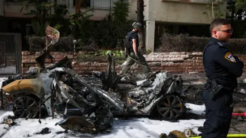 Reuters Uniformed member of Israeli security services stands in the right of the picture looking right. Beside him is the wreckage of a vehicle and behind it another man is walking.