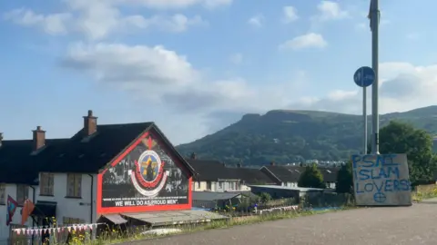 Blue spray paint on a wooden board rests against a lamppost and reads 'PSNI ISLAM LOVERS'. Behind is a mural on a terrace house that cannot be made out. 